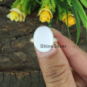 May include: A silver ring with a large, oval, white stone. The ring has a simple band and the text "ShineSilver" is visible on the stone. The ring is held up against a background of wood and yellow flowers.