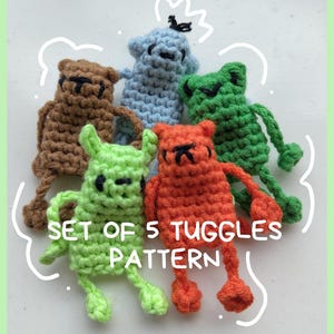 May include: A set of five small, crocheted animal-shaped toggles in brown, light blue, green, lime green, and orange. Each figure features simple stitched facial details and dangling limbs. The text SET OF 5 TUGGLES PATTERN is printed in white.