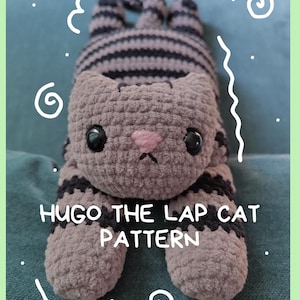 May include: A gray and black striped crocheted cat plush toy with large black eyes and a pink stitched nose. The text HUGO THE LAP CAT PATTERN is printed in white across the center of the cat, surrounded by white hand-drawn doodle lines.