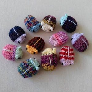 May include: A collection of 12 crocheted beetle toys in various colours, including pink, purple, blue, red, yellow, and green. The beetles are all different sizes and have different patterns.