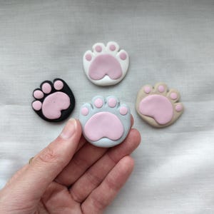May include: Four colorful cat paw-shaped decorative items. The paws are in black, white, light blue, and tan, each with a pink paw pad. These items are likely used for crafts or as decorative accents.