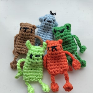May include: A collection of five small, handmade crochet figures in brown, light blue, green, lime green, and orange. Each figure features a simple rectangular body, thin dangling arms and legs, and embroidered black thread facial features.