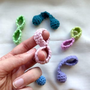 May include: A selection of small, handmade crochet pieces in various colours. The image shows abstract shapes in pink, green, blue, and purple. One pink piece is held between fingers. These items are likely decorative or for crafting purposes.
