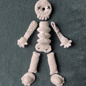 May include: A white crochet skeleton displayed on a teal background. The skeleton is constructed from individual pieces: a skull with 'x' eyes, a rib cage, arms, legs, and feet. The crochet has a textured finish.