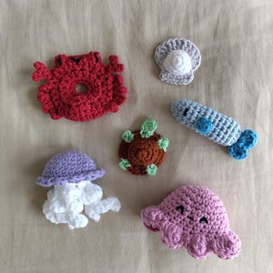 May include: A selection of handmade crochet sea creatures, including a red crab, a grey seashell, a blue fish, a brown turtle, a purple jellyfish, and a pink octopus. These soft, colourful toys are ideal for children.