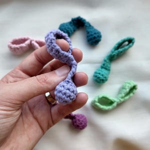 May include: Several handmade crochet items in shades of purple, pink, green, and teal. Each piece features a loop and a rounded end, possibly for decoration or embellishment. The crochet items are displayed on a light-coloured surface, showcasing the different colours.