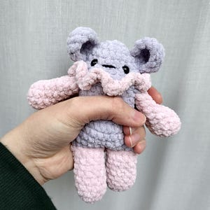 May include: A small, handmade crochet plush toy featuring a lavender-coloured head and torso with rounded ears, accented by a soft pink ruffled collar, arms, and legs. The creature has two small, round black eyes and a simple horizontal black stitched mouth.