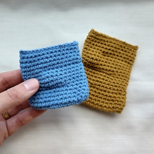 May include: Two small, handmade crochet pouches. One is held by a hand and is a light blue colour. The other pouch is a mustard yellow colour. Both pouches are rectangular and have a textured appearance due to the crochet stitch.