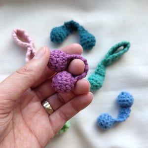 May include: A selection of small, crocheted decorative items in shades of purple, teal, pink, and blue. One purple item is held in a hand. The items feature a loop and two small spheres, suitable for embellishments or crafts.