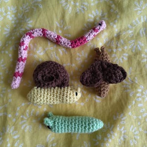 May include: A selection of handmade crochet creatures, including a pink and white snake, a brown snail, a yellow caterpillar, a green caterpillar, and a brown moth. The items are arranged on a yellow floral patterned fabric.