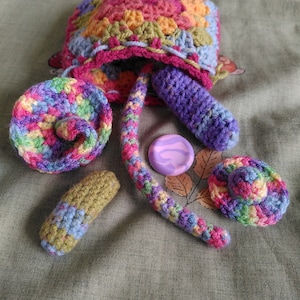 May include: A crocheted bag with a rainbow, purple, and green colour scheme. The bag is filled with crocheted items, including a small hat, a long tail, and a small rectangle. There is also a purple eraser inside the bag.