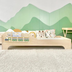 May include: A light wood children's bed with a train design on the headboard. The room features a green mountain mural, a matching dresser, and a small desk with a chair. Decorative pillows and a teddy bear are on the bed.