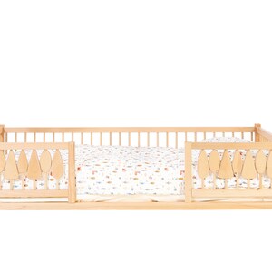 Puede incluir: Una cama de madera para ni&ntilde;os peque&ntilde;os con una s&aacute;bana estampada en blanco y marr&oacute;n. La cama tiene una barandilla de seguridad en ambos lados con recortes en forma de hoja.