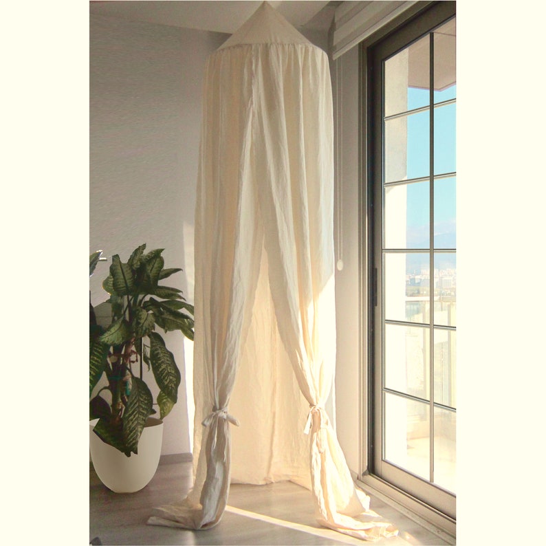 K&ouml;nnte beinhalten: Ein wei&szlig;er Baldachin aus Leinen, der von der Decke in einem Raum mit einem Fenster h&auml;ngt. Der Baldachin ist unten mit wei&szlig;en Stoffb&auml;ndern gebunden.