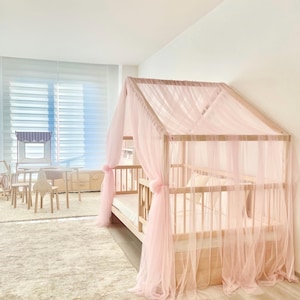 Op de afbeelding: Een licht houten kinderbed met een roze tule hemel. Het bed heeft een latten hoofdbord en voeteneind en een matras. De hemel is over het bed gedrapeerd en heeft een geplooide rand.