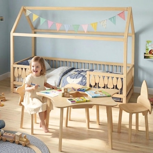 Montessori wooden house floor bed with safety railings and tree-shaped details, placed in a bright children’s room. A toddler sits at a wooden kids table drawing, with toys and books around, creating a warm, playful, and organized learning environment.