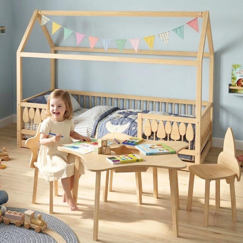 Montessori wooden house floor bed with safety railings and tree-shaped details, placed in a bright children’s room. A toddler sits at a wooden kids table drawing, with toys and books around, creating a warm, playful, and organized learning environment.