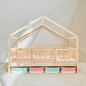 May include: A wooden house-shaped bed frame with a white mattress and a light pink and blue storage bins underneath. The bed frame has a slatted design and a decorative cutout of trees on the headboard.