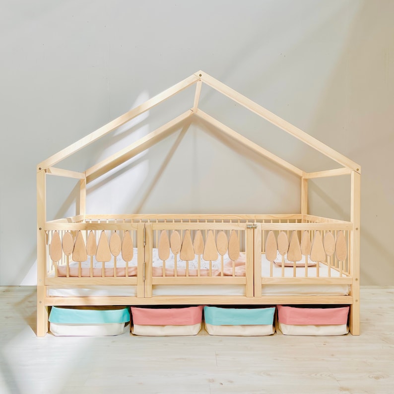 May include: A wooden house-shaped bed frame with a white mattress and a light pink and blue storage bins underneath. The bed frame has a slatted design and a decorative cutout of trees on the headboard.