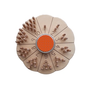 Puede incluir: Un juego de rueda de números de madera con una base circular y diez radios. Cada radio tiene un número del 1 al 10 y una fila de clavijas de madera. El centro de la rueda tiene una pegatina redonda naranja y blanca con lunares.