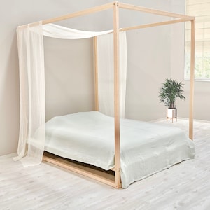 May include: A wooden canopy bed frame with a white sheer canopy and a light green bedspread. The bed is on a light wood floor.