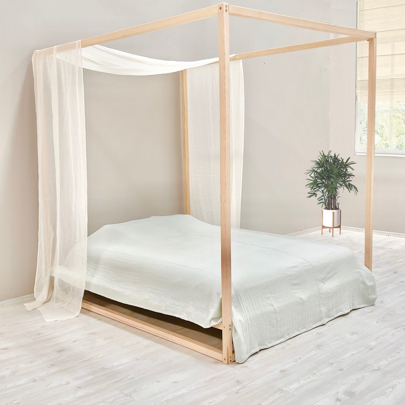 May include: A wooden canopy bed frame with a white sheer canopy and a light green bedspread. The bed is on a light wood floor.