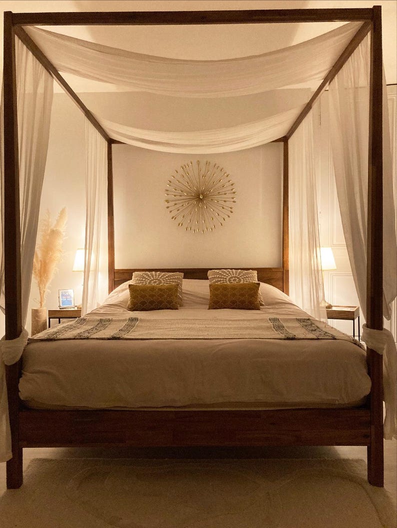 May include: A wooden four-poster bed with white sheer curtains and a white and gray patterned duvet. The bed has two pillows with gold patterned covers. A gold sunburst wall decor hangs above the bed.