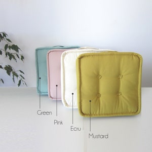 May include: Four square floor cushions in different colors: green, pink, ecru, and mustard. The cushions are made of a soft, textured fabric and have a simple, elegant design.