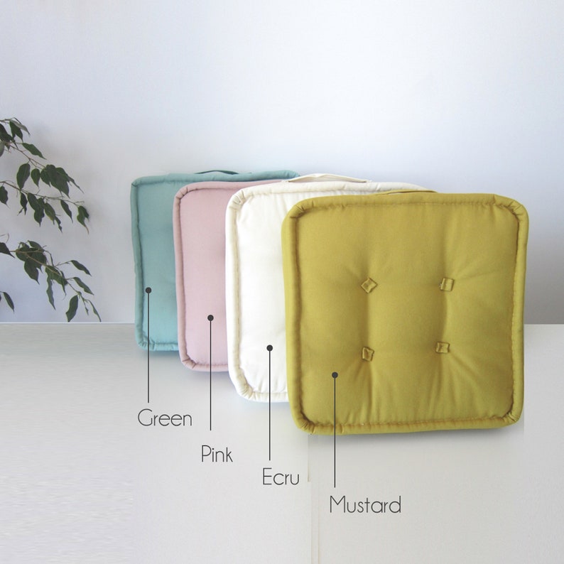 May include: Four square floor cushions in different colors: green, pink, ecru, and mustard. The cushions are made of a soft, textured fabric and have a simple, elegant design.