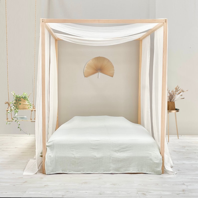 May include: A light green linen bed sheet with a white canopy draped over a wooden bed frame. The bed is in a minimalist bedroom with a white wall and a wooden fan on the wall.