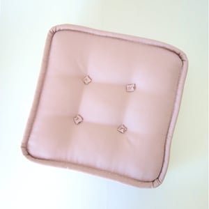 May include: A light pink square cushion with four button tufts. The cushion is made of a soft, plush fabric and has a simple, elegant design.