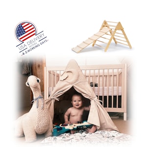 May include: A beige teepee tent with a crescent moon design on the top, set up in a nursery with a white crib and a stuffed camel toy. A baby is sitting inside the teepee playing with a toy.