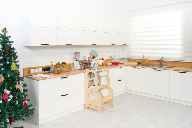 May include: A young child in a green and white chef's hat stands on a wooden learning tower in a white kitchen. The child is reaching up to a blender on the counter. There are various kitchen supplies on the counter, including a bowl of fruit, a rolling pin, and a glass jar.