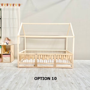 May include: A wooden toddler bed frame in the shape of a house with a white railing and a removable gate. The bed frame is made of natural wood and has a simple design. The bed frame is labeled "OPTION 10".