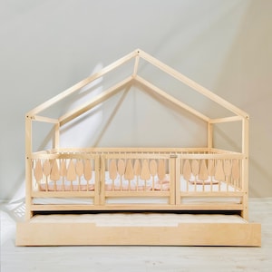 May include: A wooden toddler bed with a house-shaped frame and a pull-out drawer. The bed has a white mattress and a wooden railing with decorative cutouts.
