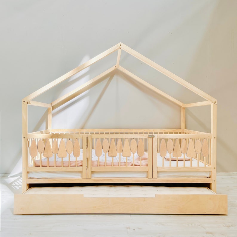 May include: A wooden toddler bed with a house-shaped frame and a pull-out drawer. The bed has a white mattress and a wooden railing with decorative cutouts.