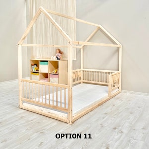 May include: A wooden house-shaped bed frame with a white mattress and a safety rail. The bed frame has a small shelf unit on the side with a doll and toys inside. The text "OPTION 11" is printed on the floor in front of the bed.