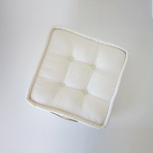May include: A white, square cushion with a tufted design and button accents. The cushion is made of a soft, plush fabric.