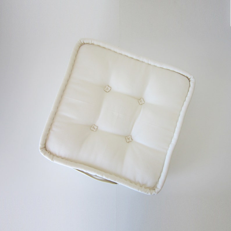 May include: A white, square cushion with a tufted design and button accents. The cushion is made of a soft, plush fabric.