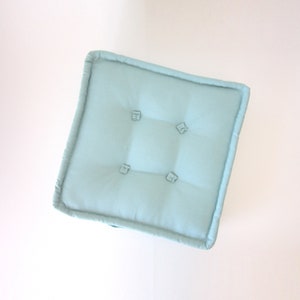 May include: A light blue square cushion with four button tufts. The cushion is made of a soft, textured fabric.