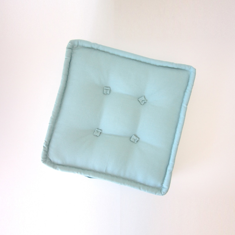 May include: A light blue square cushion with four button tufts. The cushion is made of a soft, textured fabric.