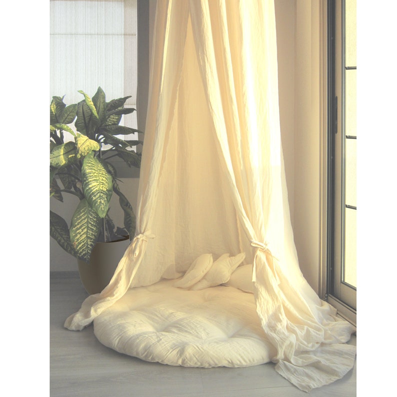 K&ouml;nnte beinhalten: Ein wei&szlig;es Baldachinbett aus Baumwolle mit einem wei&szlig;en Kissen und Kissen. Das Bett steht in einem Raum mit einem Fenster und einer Pflanze.