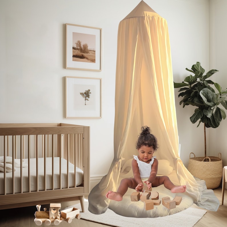 K&ouml;nnte beinhalten: Ein beigefarbenes Baldachin-Zelt mit einem wei&szlig;en R&uuml;schenrand am Boden. Ein junges M&auml;dchen sitzt im Zelt und spielt mit Holzkl&ouml;tzen. Das Zelt ist in einem Kinderzimmer mit einem Kinderbett und gerahmten Bildern an der Wand aufgebaut.