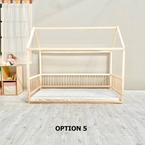 May include: A wooden house-shaped bed frame with a white mattress. The bed frame has a slatted headboard and footboard, and a railing around the sides. The bed frame is on a light wood floor. The text "OPTION 5" is printed below the bed frame.
