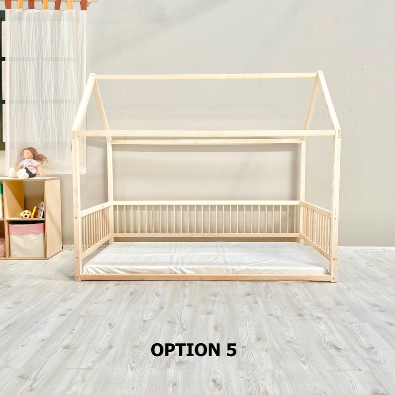 May include: A wooden house-shaped bed frame with a white mattress. The bed frame has a slatted headboard and footboard, and a railing around the sides. The bed frame is on a light wood floor. The text "OPTION 5" is printed below the bed frame.