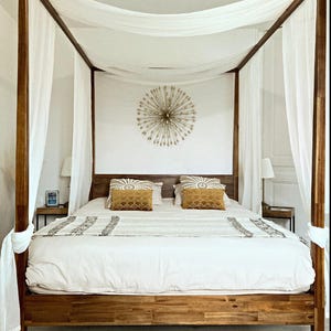 May include: A wooden canopy bed with white sheer curtains and a white duvet. The bed has two yellow throw pillows and a gold starburst wall decor.