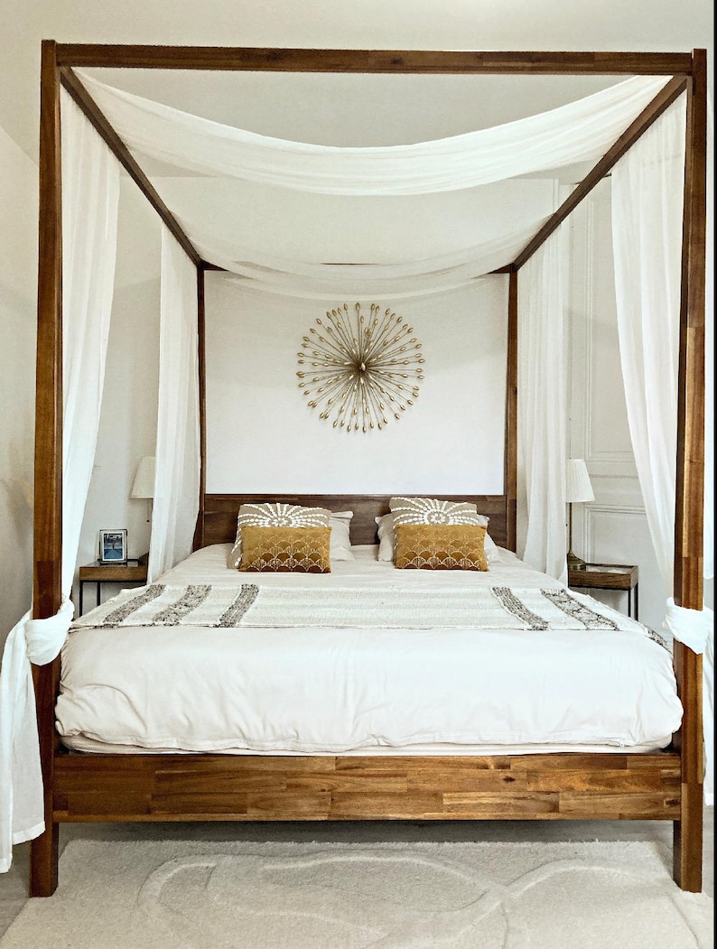 May include: A wooden canopy bed with white sheer curtains and a white duvet. The bed has two yellow throw pillows and a gold starburst wall decor.