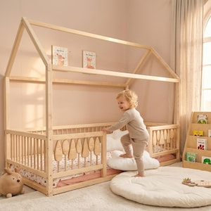 Montessori house floor bed made from natural wood with safety railings and decorative tree-shaped slats, placed in a soft-toned children’s room. A toddler is climbing into the bed using cushions, with bookshelves, toys, and warm natural light creating a cozy, child-friendly environment.