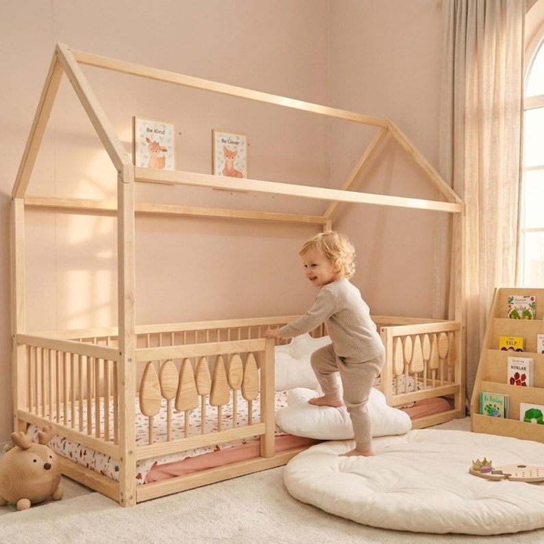 Montessori house floor bed made from natural wood with safety railings and decorative tree-shaped slats, placed in a soft-toned children’s room. A toddler is climbing into the bed using cushions, with bookshelves, toys, and warm natural light creating a cozy, child-friendly environment.