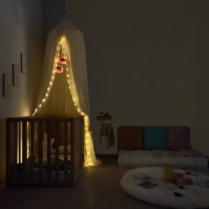 K&ouml;nnte beinhalten: Ein wei&szlig;er Baldachin mit Lichterketten h&auml;ngt &uuml;ber einem Holzbettchen. Das Bettchen steht neben einem wei&szlig;en Futon mit Kissen und einem wei&szlig;en Teppich. Der Raum ist schwach beleuchtet.
